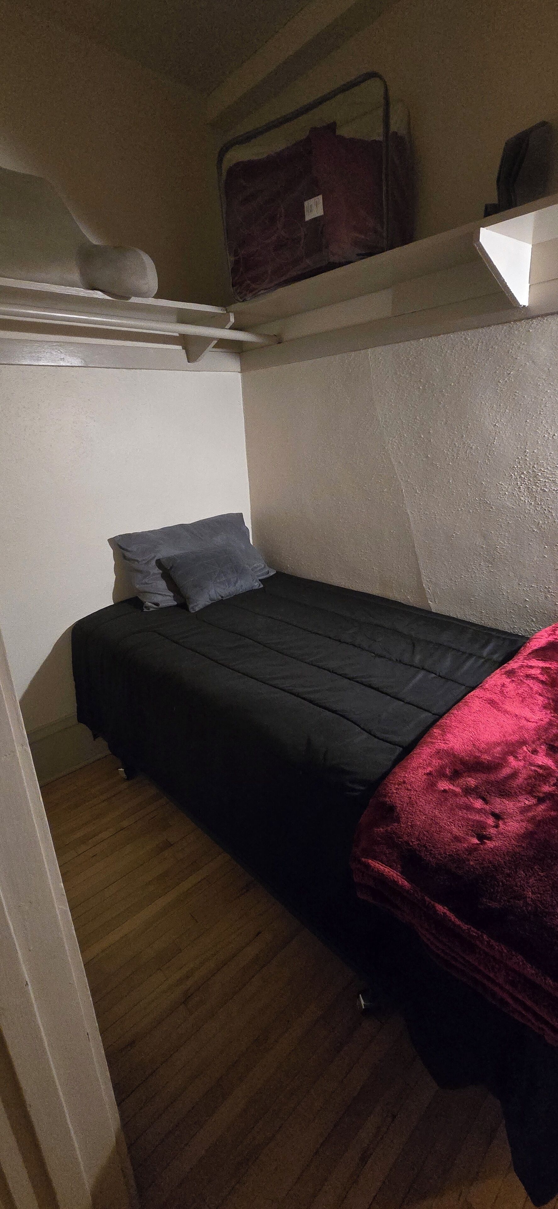 A small, dimly lit room with a single bed, a red blanket, and a shelf with bags.