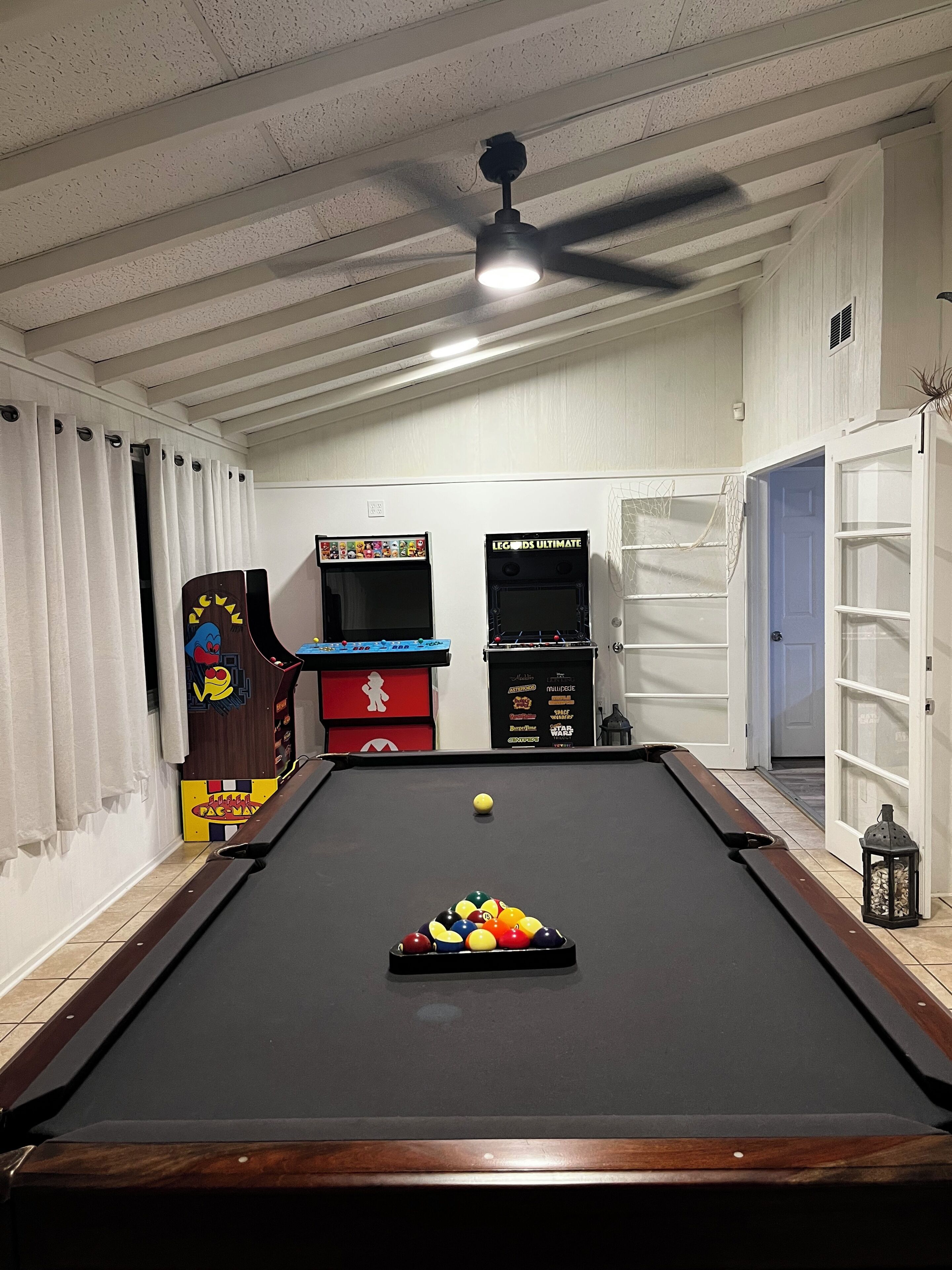 Une table de billard avec une bille en jeu, des jeux d’arcade et un ventilateur de plafond dans une pièce aux murs blancs et dotée d’une porte.