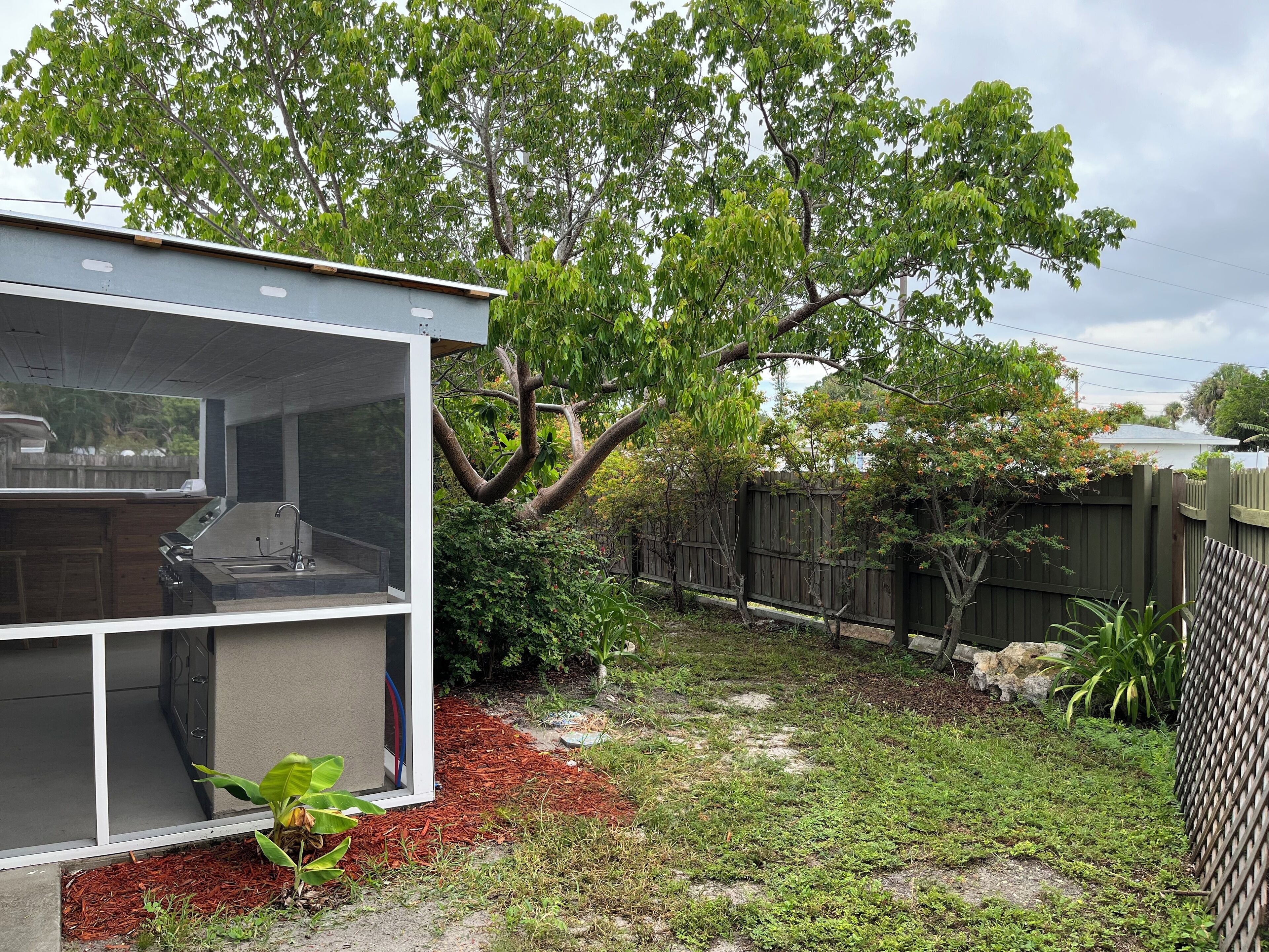 A modern house with a covered outdoor kitchen area, a fenced backyard, and a large tree providing shade.