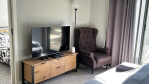 A modern hotel room with a large flat-screen TV, a wooden side table, a dark armchair, a sofa, and a floor lamp. - A Little Southern Comfort - granny flat in Sanctuary Point  (Sanctuary Point)