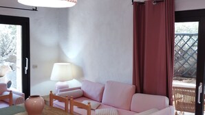 Quarto com sofá rosa, mesa de jantar de madeira, cadeiras, luminária pendente e janela com cortinas.