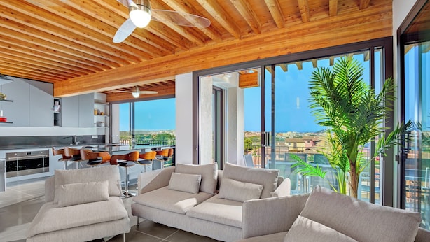 Una sala de estar moderna con un sofá grande, un área de comedor con taburetes altos y una cocina. La habitación tiene ventanas amplias que ofrecen vistas de la ciudad.