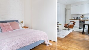 A bedroom with a bed, a nightstand, a lamp, and a chair. The room has wooden flooring and white walls. - Le Plaisir du Pantheon + Seine Cruise (Aug only!) (Paris)