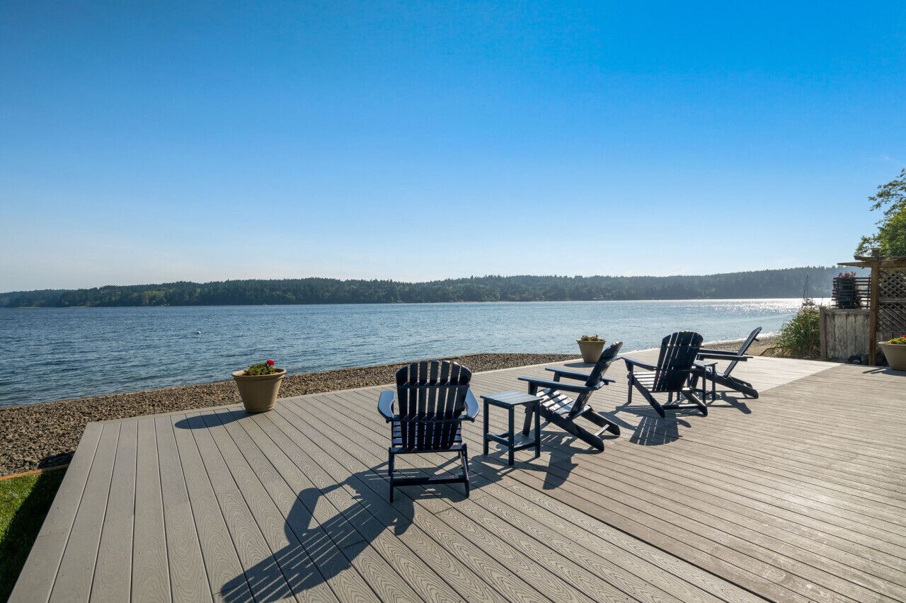 Um deck de madeira com cadeiras Adirondack e uma pequena mesa, com vista para um lago calmo e uma margem florestada.