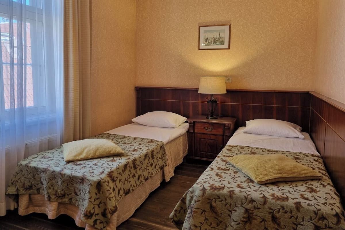 Une chambre d’hôtel avec deux lits simples, une table de chevet avec une lampe, un tableau encadré au mur et une fenêtre avec des rideaux légers.