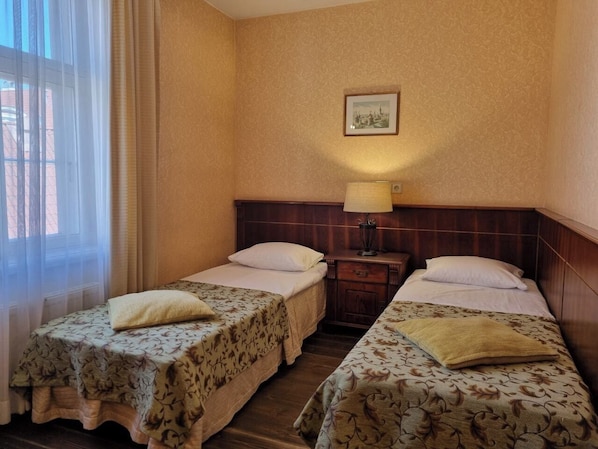 Ein Hotelzimmer mit zwei Einzelbetten, einem Nachttisch mit Lampe, einem gerahmten Bild an der Wand und einem Fenster mit durchsichtigen Vorhängen.
