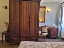 A bedroom with a large wooden wardrobe, a chair, a desk with a lamp, and a bed with a floral bedspread.