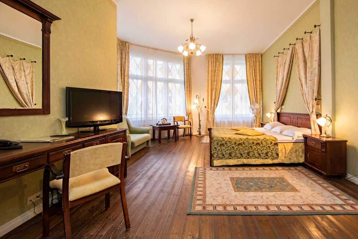 Une chambre d’hôtel avec un plancher en bois, un lit recouvert d’une couvre-lit à motifs floraux, un bureau avec une télévision, une chaise et une table de chevet avec une lampe.