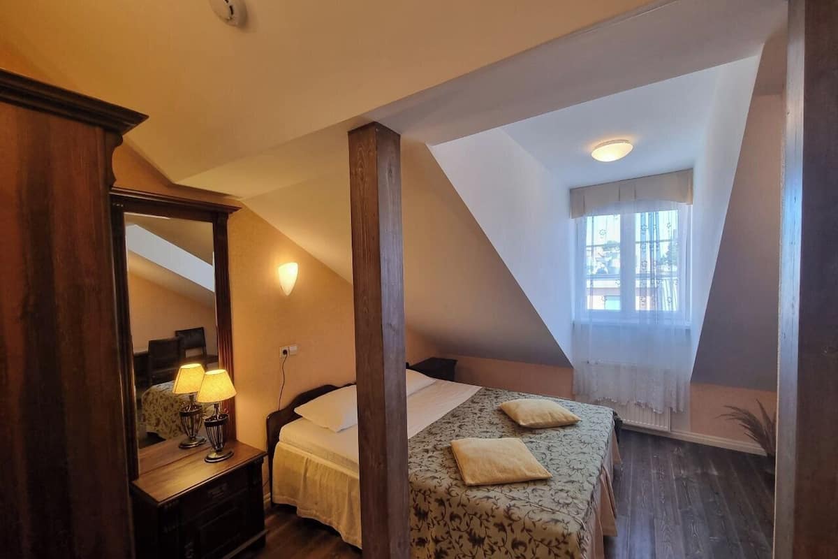 Une chambre avec un plafond en pente, un grand miroir, un lit recouvert d’une couvre-lit à motifs floraux et une fenêtre ornée de rideaux légers.