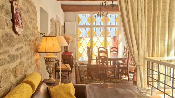 Ein gemütliches Wohnzimmer mit einem braunen Ledersofa, einem Tisch mit einer Lampe und Blick ins Freie durch ein Fenster mit gemusterten Vorhängen.