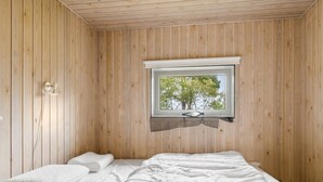 Een kleine kamer met houten lambrisering, een enkel bed, een raam met uitzicht op bomen en een eenvoudig nachtkastje.