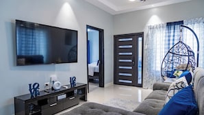 Una sala de estar moderna con un televisor de pantalla plana, un mueble negro para equipo multimedia, un sofá gris con cojines azules, una silla colgante y una puerta que da a otra habitación.