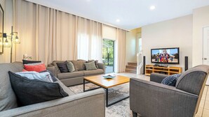 Een moderne woonkamer met een bank, fauteuil en salontafel. Een televisie is aan de muur bevestigd en er is een raam met gordijnen.