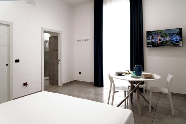A hotel room with a bed, a dining table with chairs, a TV mounted on the wall, and a bathroom visible through an open door.