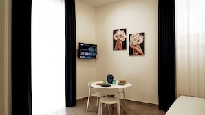 Un pequeño comedor con una mesa y sillas blancas, un televisor de pantalla plana montado en la pared y dos cuadros con motivos florales.