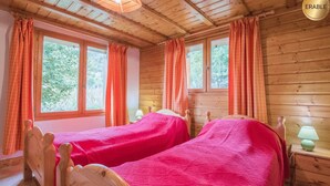 A wooden cabin with two beds, each with a pink bedspread, and orange curtains. There are wooden headboards and a small table with a lamp.
