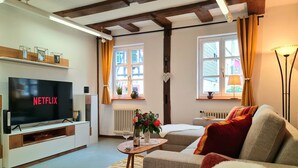 Ein gemütliches Wohnzimmer mit einem Flachbildfernseher, der 'NETFLIX' zeigt, einer Couch, einem Couchtisch mit Blumen und einer Vase sowie einem Fenster mit Vorhängen.