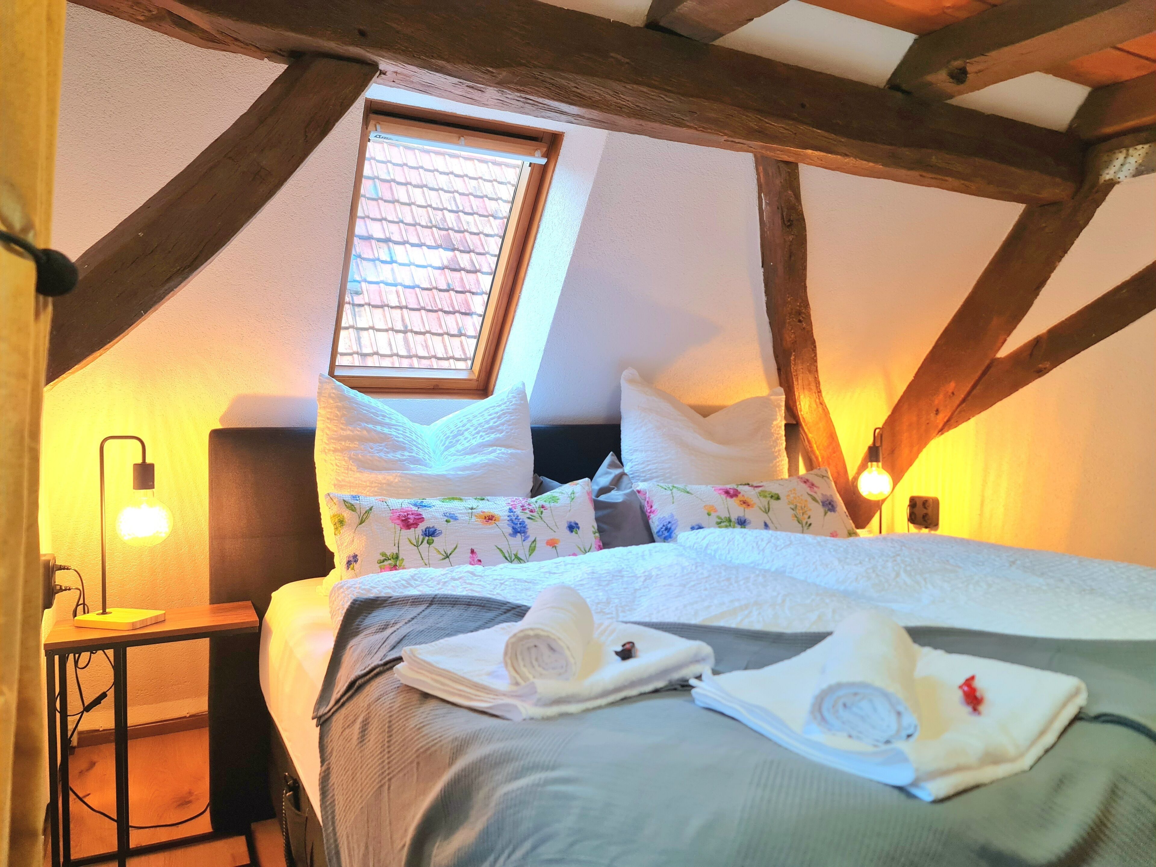 A cozy attic bedroom with a sloped ceiling, wooden beams, a skylight, a bed with floral pillows, and a bedside table with a lamp.