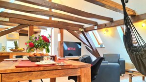 Ein gemütliches Wohnzimmer mit einem Holzesstisch, einer bequemen Couch und einer Hängematte. Ein Fernseher mit dem Schriftzug 'NETFLIX' ist an der Wand befestigt.