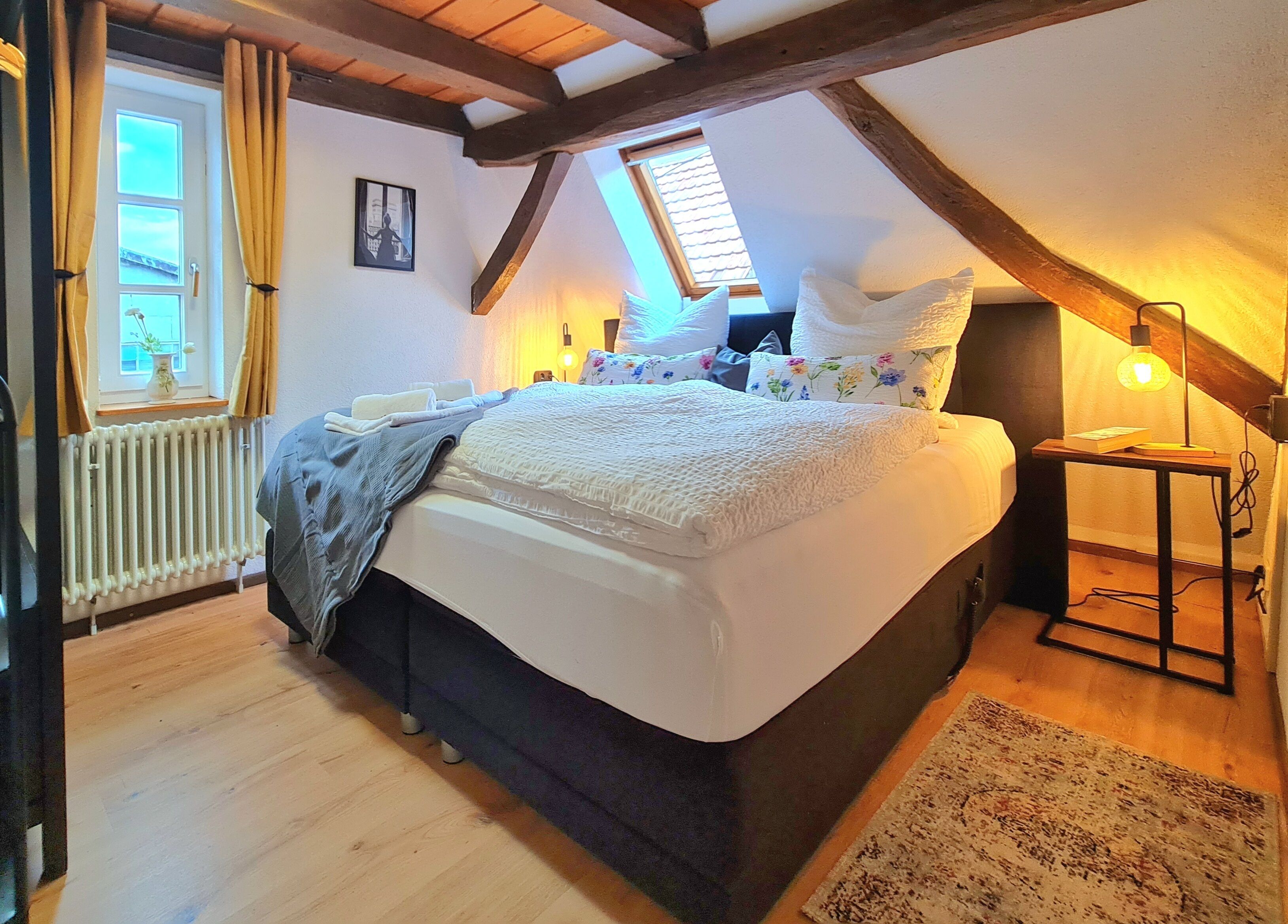A bedroom with a sloped ceiling, a bed with white bedding, a bedside table with a lamp, and a window with curtains.