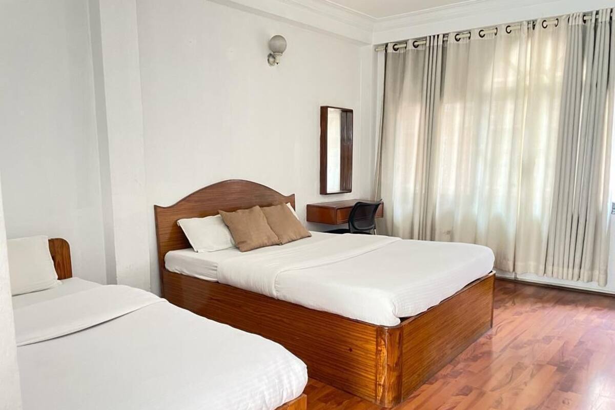 Una habitación de hotel con dos camas, suelo de madera, un escritorio con una silla, un espejo y una ventana con cortinas.