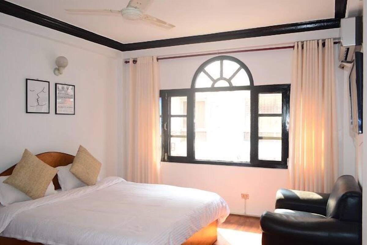 Una habitación de hotel con cama, una silla, ventilador de techo, ventana con cortinas y decoración en la pared.