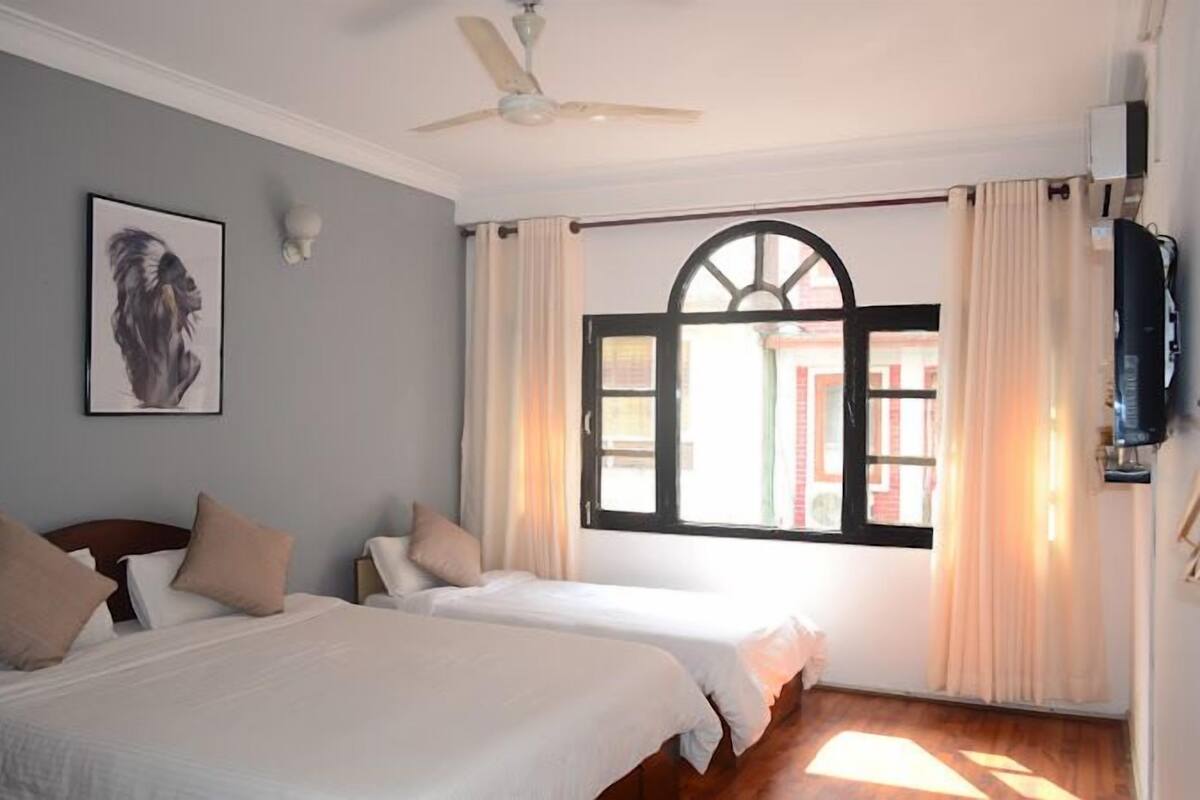 Una habitación de hotel con dos camas, un ventilador de techo, un ventanal grande con cortinas y un televisor montado en la pared.