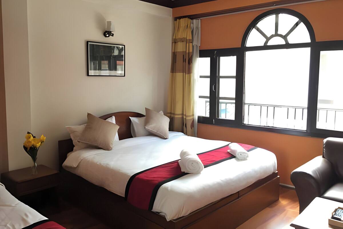 Una habitación de hotel con una cama grande, una mesita de noche con un florero y una ventana con vistas al exterior, además de una silla.