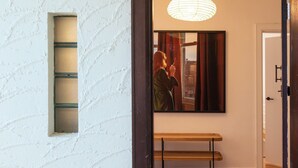 A hallway with a textured white wall, a window with a grid pattern, a wooden bench, and a framed painting of a person holding a book. - Pet Friendly - Charnwood Charm in St Kilda (St Kilda)