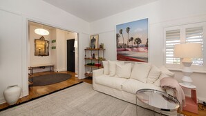 A living room with a white sofa, a glass coffee table, a painting of palm trees, and a bookshelf. - Pet Friendly - Charnwood Charm in St Kilda (St Kilda)