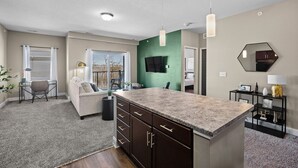 A modern kitchen with a central island, a dining area with a sofa and table, and a living room with a TV and green accent wall. - Top Floor, Modern & Upgraded with Open Balcony (West Des Moines)