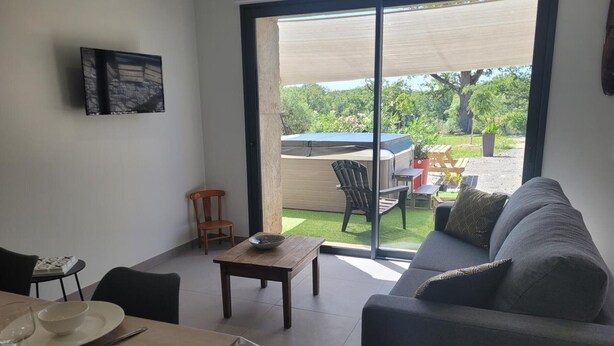 A modern living room with a sofa, a dining table with a bowl and wine glass, and a sliding glass door leading to an outdoor patio with a hot tub and garden furniture. - Peaceful cottage with spa and air conditioning in Couzon (Couzon)