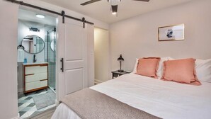 Un dormitorio con una cama, una mesita de noche, una lámpara, un ventilador de techo, una puerta que da al baño y un cuadro en la pared.