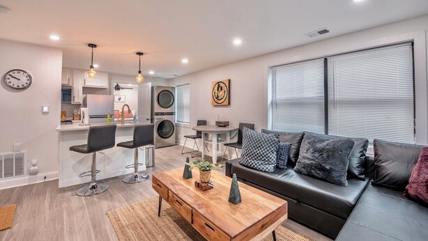 Una sala de estar moderna con un sofá, una mesa de centro y sillas. Hay una zona de cocina con electrodomésticos y una mesa de comedor.