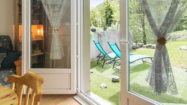 A room with a sliding glass door leading to a patio with a lawn, two blue lounge chairs, and a decorative curtain. - Cozy 1-Bed Apartment for 4 with Garden, Parking, Near Cauterets Center and Thermal Spa (Cauterets)