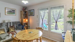 A cozy living room with a wooden dining table, a sofa with patterned cushions, a bookshelf, and a large window with curtains. - Cozy 1-Bed Apartment for 4 with Garden, Parking, Near Cauterets Center and Thermal Spa (Cauterets)