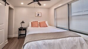 Un dormitorio con cama, mesita de noche, ventilador de techo, ventana con persianas y un cuadro en la pared.