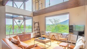 Un salón amplio con ventanas grandes, suelo de madera, un sofá de cuero, una mesa de centro y un televisor.