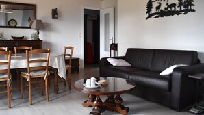 A living room with a black leather sofa, wooden dining chairs, a round coffee table, and a wall art depicting a mountain scene.
