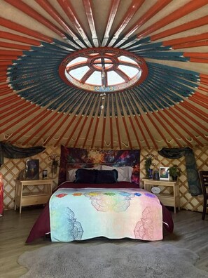 A yurt interior with a bed, a circular skylight, and a patterned tapestry.