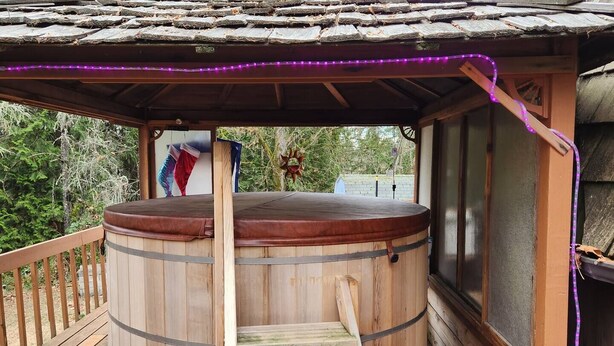 A wooden hot tub with a wooden ladder, surrounded by a wooden deck and a covered area with a roof.