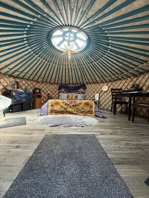 A yurt interior with a central circular skylight, a bed with a colorful blanket, and a dining area with wooden furniture.