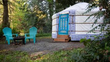 Une yourte avec une porte bleue, deux chaises turquoise et une table, le tout situé dans un cadre forestier.