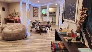 A cozy room with a large bean bag, a dining table with chairs, a small kitchen area, and a wall with decorative items.