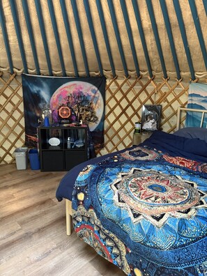 Una habitación acogedora con una cama, un tapiz en la pared, una mesita con objetos y suelo de madera.