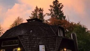 A wooden cabin with a porch, surrounded by trees, under a sunset sky.