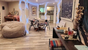Una sala de estar moderna con un sillón tipo bean bag grande, una mesa de comedor con sillas, una cocina al fondo y elementos decorativos como un cuadro enmarcado y un jarrón con flores secas.