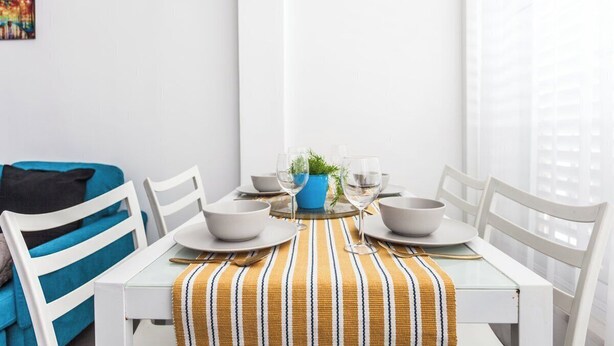 Un coin repas avec une table blanche dressée pour deux personnes, un vase bleu orné de verdure et une nappe à rayures.