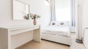 A bedroom with a bed, a bedside table, a mirror, a vase with flowers, and a window with curtains. - Port 2 Bedroom with 2 showers apartment (Tel Aviv)
