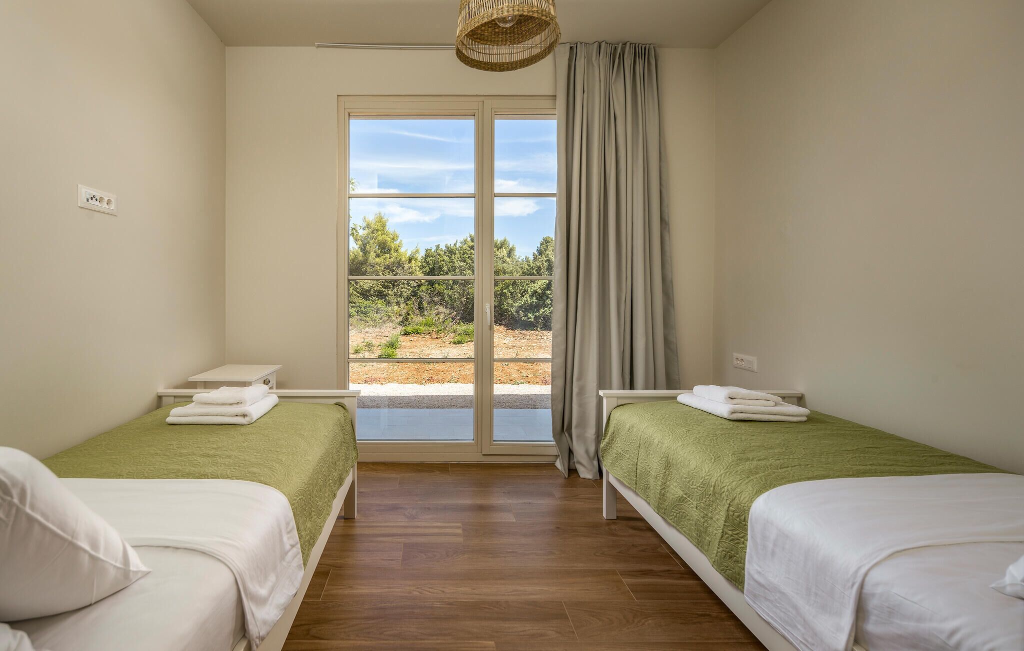 Ein Zimmer mit zwei Betten, einem Fenster mit Blick auf Bäume und einer Lampenschirm aus Weidengeflecht.